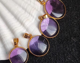 May include: Four round pendants with a gold-colored frame and bail. Each pendant features a deep purple, reflective stone. The pendants are arranged on a white seashell and a black surface.