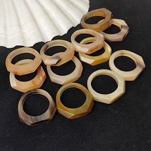 Op de afbeelding: Een verzameling edelsteenringen in verschillende tinten bruin, beige en crème. De ringen hebben een geometrisch, facetgeslepen ontwerp. Op de achtergrond een witte zeeschelp, op een zwart oppervlak.
