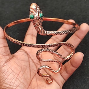 Può includere: Un braccialetto di serpente in rame con occhi di pietre preziose verdi. Il serpente è avvolto e il braccialetto è aperto.