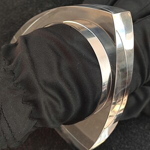 Op de afbeelding: Een gepolijste zilverkleurige manchetarmband met een geometrisch ontwerp. De armband heeft een brede, gebogen vorm met een platte bovenkant en een glad, reflecterend oppervlak. De armband wordt gedragen op een zwarte stoffen achtergrond.