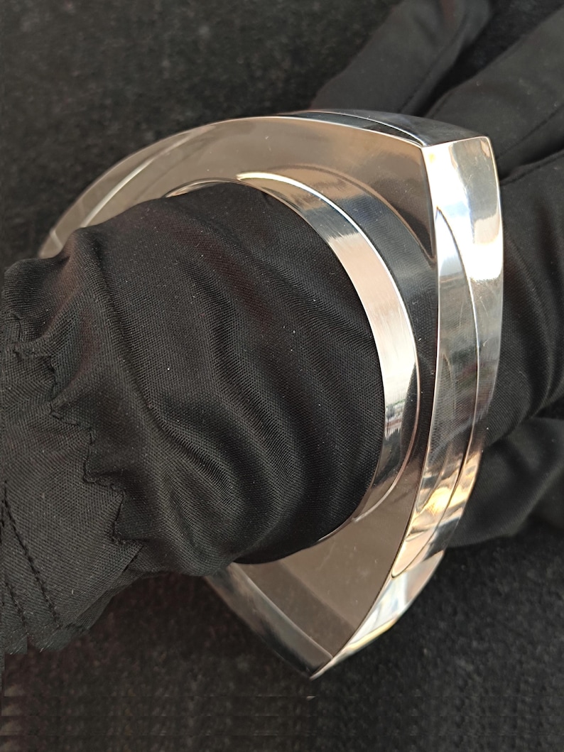 Op de afbeelding: Een gepolijste zilverkleurige manchetarmband met een geometrisch ontwerp. De armband heeft een brede, gebogen vorm met een platte bovenkant en een glad, reflecterend oppervlak. De armband wordt gedragen op een zwarte stoffen achtergrond.
