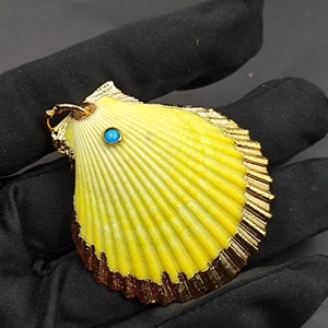 K&ouml;nnte beinhalten: Ein gelber Muschel-Anh&auml;nger mit goldener Umrandung und einem kleinen t&uuml;rkisfarbenen Edelstein in der Mitte.