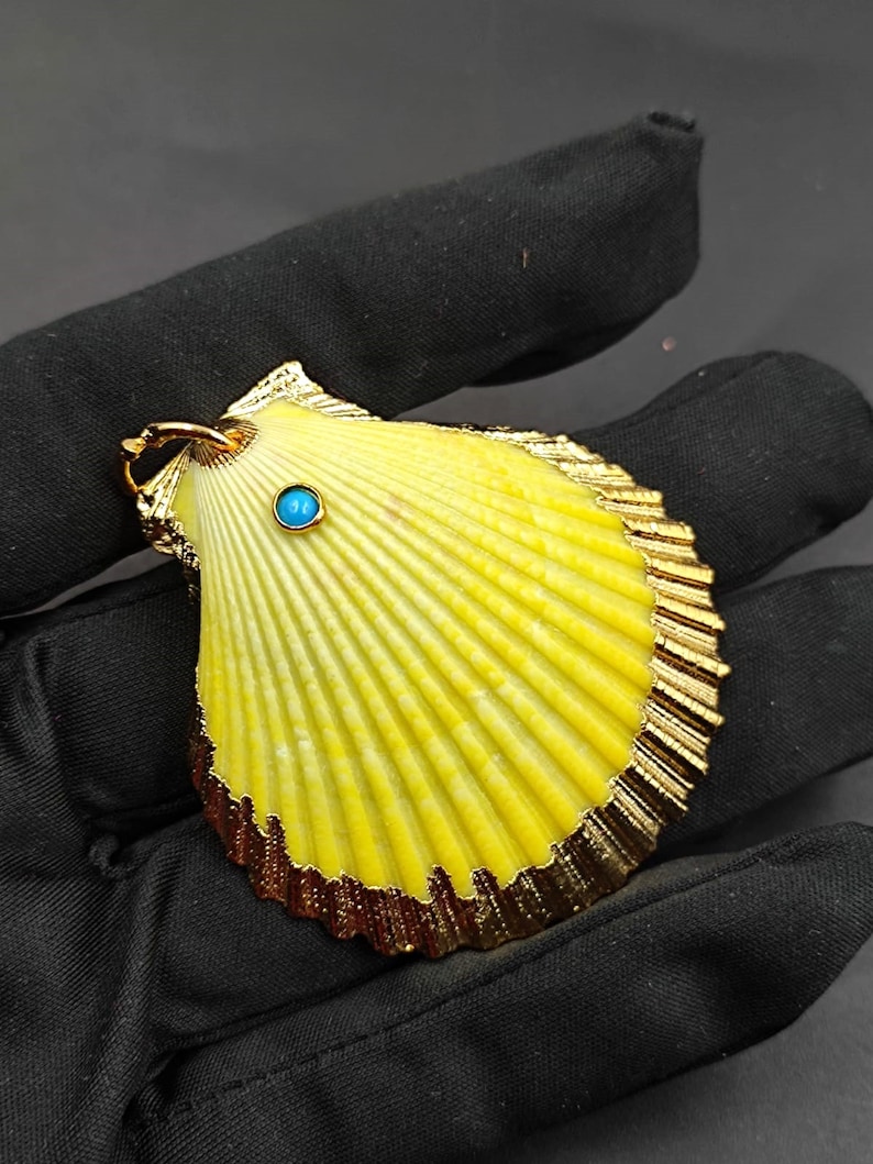 K&ouml;nnte beinhalten: Ein gelber Muschel-Anh&auml;nger mit goldener Umrandung und einem kleinen t&uuml;rkisfarbenen Edelstein in der Mitte.