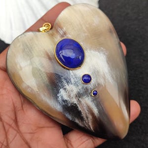 Puede incluir: Un colgante en forma de corazón hecho de cuerno marrón y blanco con una piedra preciosa azul grande y dos piedras preciosas azules más pequeñas engastadas en oro.