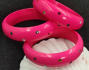 May include: Three bright pink bangles with colorful rhinestones. The bangles are stacked on top of each other, with a white seashell in the foreground. The rhinestones are in various colors, including green, red, and clear.