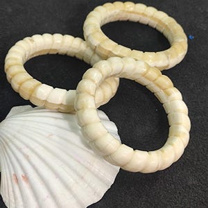 May include: Three off-white, textured bracelets with a segmented design are displayed on a dark surface, one resting on a white seashell. The bracelets have a slightly curved shape and a repeating pattern of rounded segments.