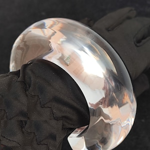 Peut inclure: Un bracelet jonc large et poli, de couleur claire. Le bracelet est de forme incurvée et pleine, avec une surface lisse et réfléchissante. La lumière se reflète sur la surface, créant un aspect brillant. Le bracelet est présenté sur un fond noir.