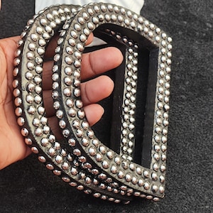 May include: Three D-shaped belt buckles with a dark gray finish and silver-colored studs. The buckles are stacked, showcasing the studded detail around the edges. The background is a dark gray fabric.