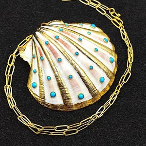 Puede incluir: Un collar de cadena dorada con un gran colgante de concha. La concha está pintada de oro y tiene detalles en azul turquesa.