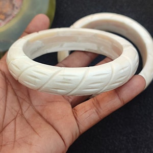 May include: Two white bone bangles with a carved leaf design. The bangles are stacked on top of each other and are being held by a hand.