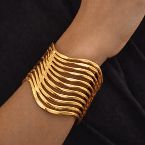 Op de afbeelding: Een goudkleurige manchetarmband met een golvend, gelaagd ontwerp. De armband is gemaakt van meerdere parallelle banden die een textuurrijke, vloeiende uitstraling creëren. Het sieraad wordt gedragen om een pols tegen een donkere achtergrond.