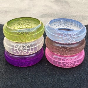 May include: Set of six colorful translucent plastic bangles with a spiderweb design. The bangles are stacked in pairs, with green and white on the left and blue and pink on the right.