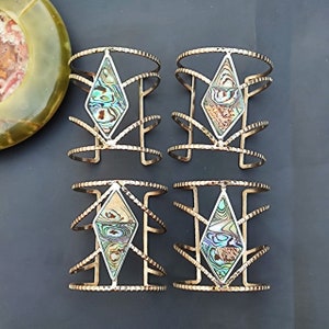 Könnte beinhalten: Vier goldfarbene Armreifen mit rautenförmiger Abalone-Muschel-Einlage. Die Armreifen haben ein strukturiertes Design und sind auf einer dunkelgrauen Oberfläche angeordnet. Die Abalone-Muscheln zeigen schillernde Farben.