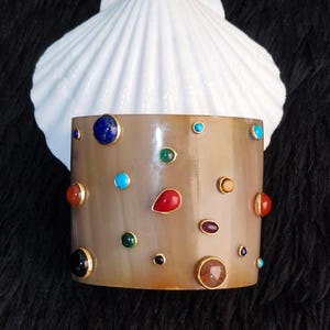 Puede incluir: Un brazalete ancho y liso de color marrón claro que presenta un surtido de pequeñas piedras preciosas de colores, redondas y en forma de lágrima, engastadas en biseles de metal dorado dispersas por toda la superficie.