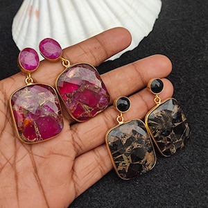 May include: Gold-toned earrings with large, rectangular, and oval gemstones. One pair features deep red and purple stones, while the other has black and brown stones. The earrings are displayed on a dark surface.