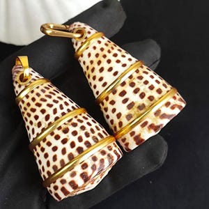 May include: Two cone-shaped pendants with a natural shell pattern of brown spots on a cream background. Each pendant is accented with gold-colored metal bands and a gold-colored bail.