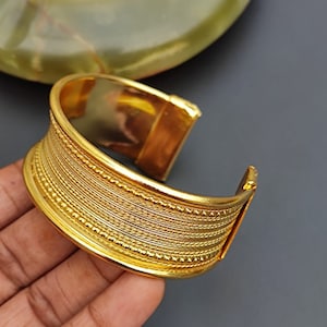 May include: A gold-toned metal bangle bracelet with a textured design. The bracelet has a series of raised lines and dots.