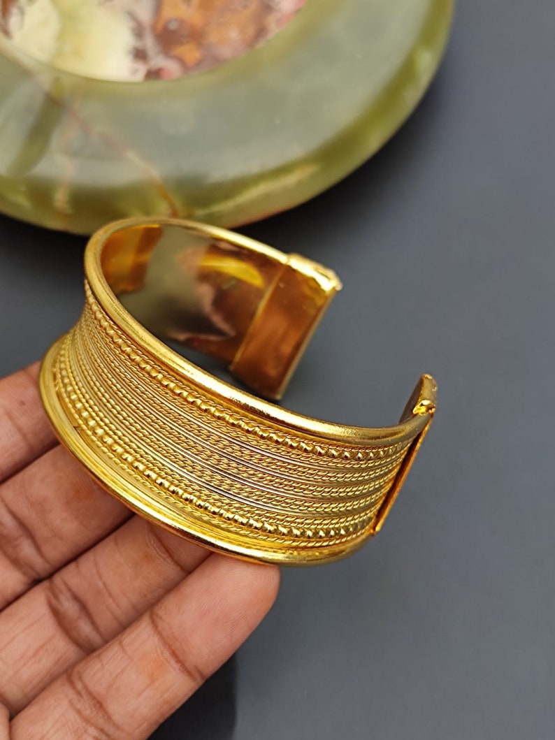 May include: A gold-toned metal bangle bracelet with a textured design. The bracelet has a series of raised lines and dots.