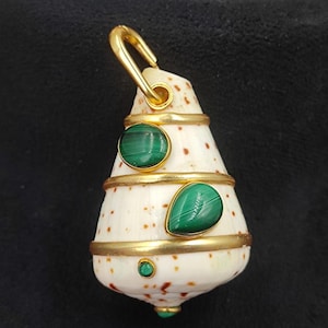 May include: A pendant featuring a white seashell with brown spots, adorned with gold accents and green malachite stones. The pendant has a gold bail for hanging.