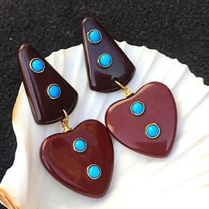 May include: A pair of dangle earrings with a geometric triangle shape and a heart shape. The earrings are made of dark brown resin and feature turquoise blue stones.