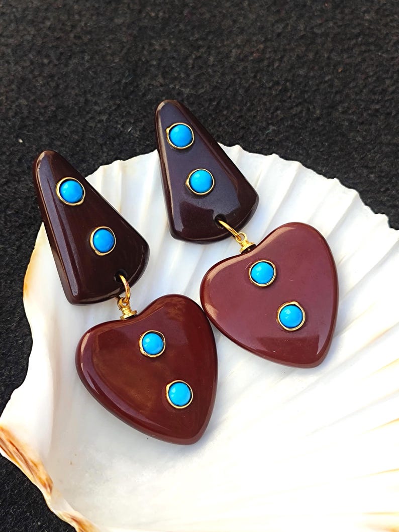 May include: A pair of dangle earrings with a geometric triangle shape and a heart shape. The earrings are made of dark brown resin and feature turquoise blue stones.
