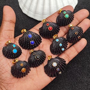 May include: A selection of black seashell pendants with gold-coloured bails, each embellished with colourful gemstones. The gemstones include blue, red, green, and yellow, providing a vibrant contrast. The pendants are displayed on a dark surface.