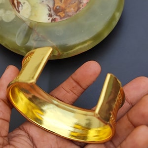 May include: A gold-toned bangle bracelet with a textured design. The bracelet is open and has a wide, flat band.