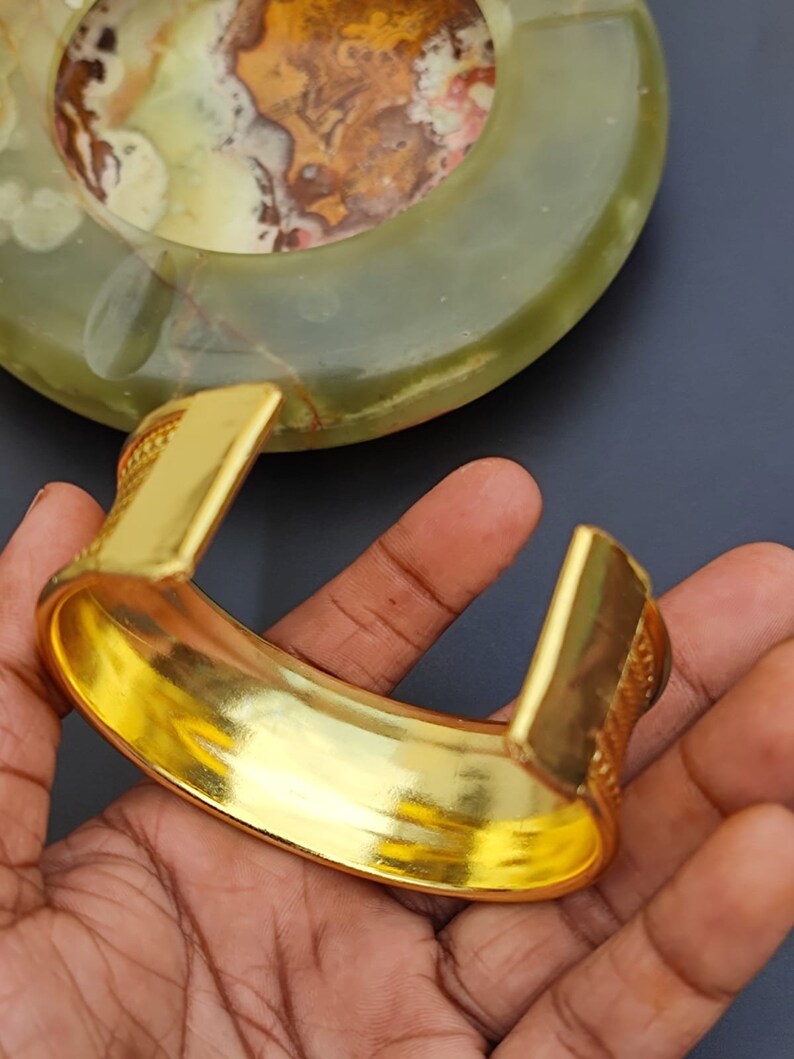 May include: A gold-toned bangle bracelet with a textured design. The bracelet is open and has a wide, flat band.