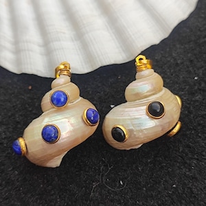 Peut inclure: Deux boucles d'oreilles en forme de coquillage avec des détails dorés et des pierres précieuses. L'une a des pierres bleues, l'autre des pierres noires. Elles sont présentées sur un coquillage blanc et un fond sombre.