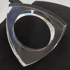Op de afbeelding: Een heldere, geometrische armband. De armband heeft een driehoekige buitenkant met een cirkelvormige opening. Het transparante materiaal laat licht door, waardoor een strakke en moderne esthetiek ontstaat. De armband wordt op een zwart oppervlak gepresenteerd.