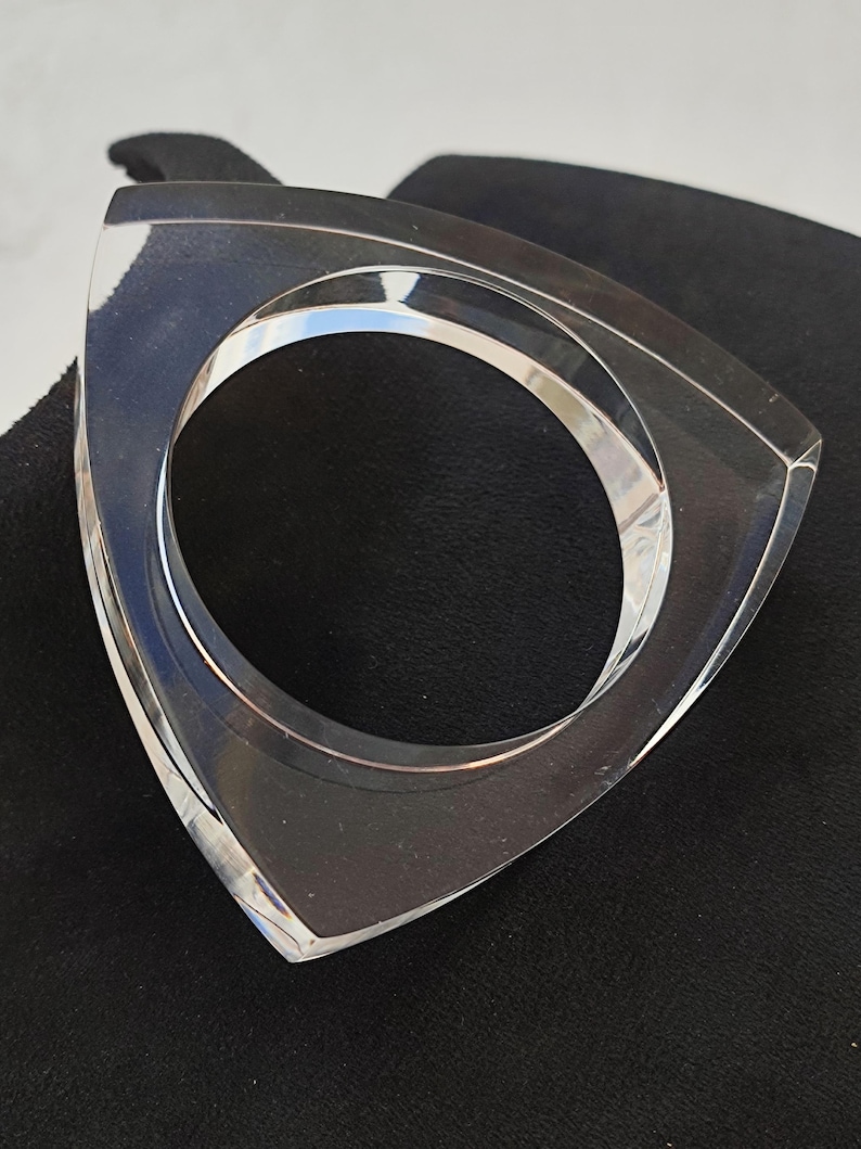 Op de afbeelding: Een heldere, geometrische armband. De armband heeft een driehoekige buitenkant met een cirkelvormige opening. Het transparante materiaal laat licht door, waardoor een strakke en moderne esthetiek ontstaat. De armband wordt op een zwart oppervlak gepresenteerd.