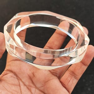 Puede incluir: Pulsera transparente de acr&iacute;lico con dise&ntilde;o geom&eacute;trico. El brazalete transparente tiene una forma facetada y angular, que refleja la luz. La pulsera se sostiene en una mano, mostrando su superficie pulida y su estilo moderno.