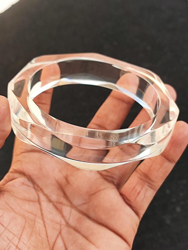 Puede incluir: Pulsera transparente de acr&iacute;lico con dise&ntilde;o geom&eacute;trico. El brazalete transparente tiene una forma facetada y angular, que refleja la luz. La pulsera se sostiene en una mano, mostrando su superficie pulida y su estilo moderno.