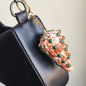 Puede incluir: Un dije decorativo de concha con patrones marrones y blancos, adornado con detalles verdes y turquesas. El dije está unido a una cadena dorada y a un bolso de cuero negro. La concha tiene forma de cono.