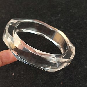 Puede incluir: Brazalete transparente y facetado. El brazalete transparente tiene un dise&ntilde;o geom&eacute;trico con m&uacute;ltiples lados, que reflejan la luz. El brazalete est&aacute; hecho de un material transparente y se muestra sobre un fondo oscuro.