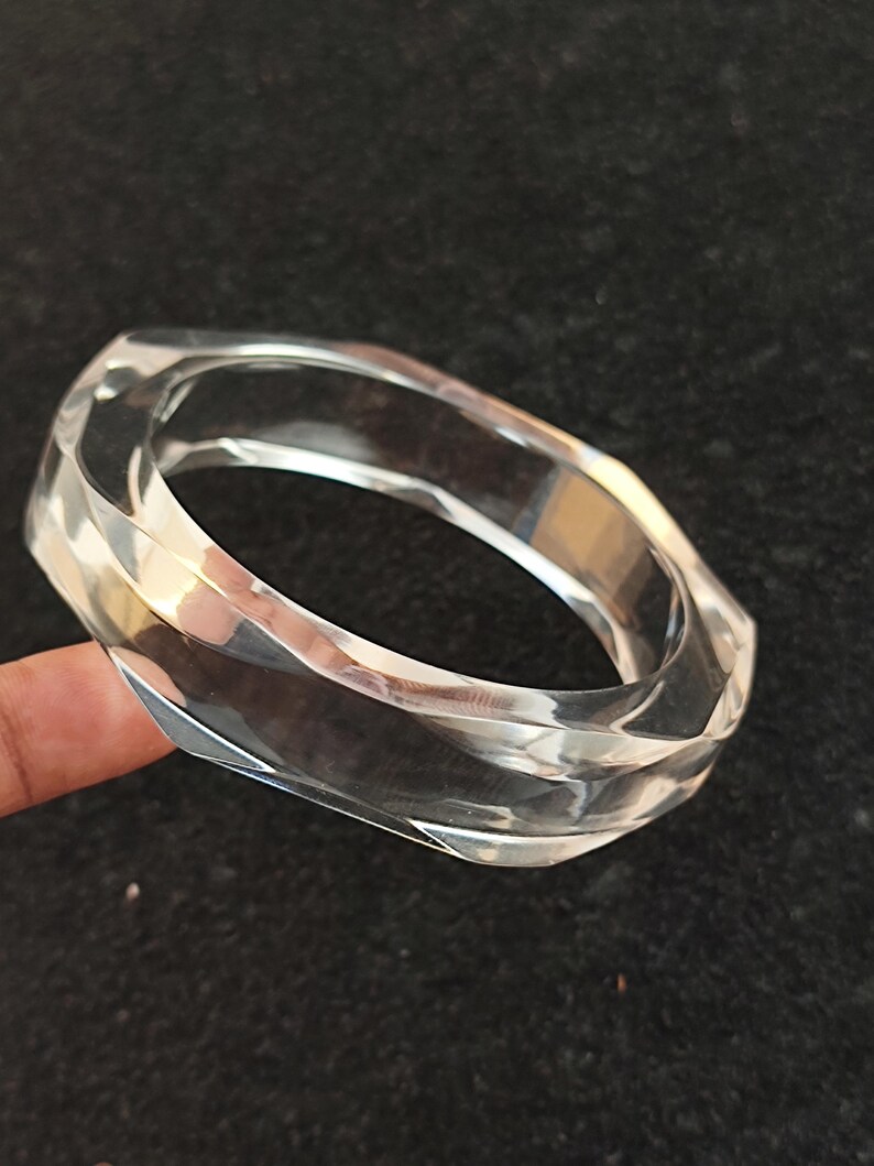 Puede incluir: Brazalete transparente y facetado. El brazalete transparente tiene un dise&ntilde;o geom&eacute;trico con m&uacute;ltiples lados, que reflejan la luz. El brazalete est&aacute; hecho de un material transparente y se muestra sobre un fondo oscuro.