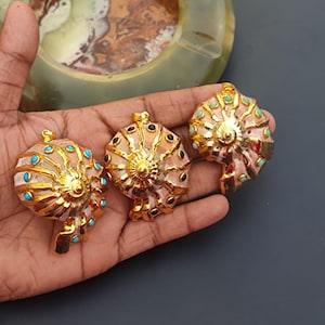 Könnte beinhalten: Drei vergoldete Muschel Anhänger mit bunten Edelstein-Akzenten. Die Muscheln sind rosa und weiß mit goldenen Akzenten. Die Edelsteine sind türkis, schwarz und grün.