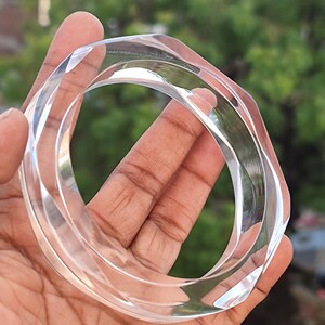 Puede incluir: Brazalete de acr&iacute;lico transparente y facetado. El brazalete transparente tiene un dise&ntilde;o geom&eacute;trico con m&uacute;ltiples lados. La luz atrapa los bordes, creando reflejos. El brazalete se sostiene en una mano sobre un fondo verde borroso.