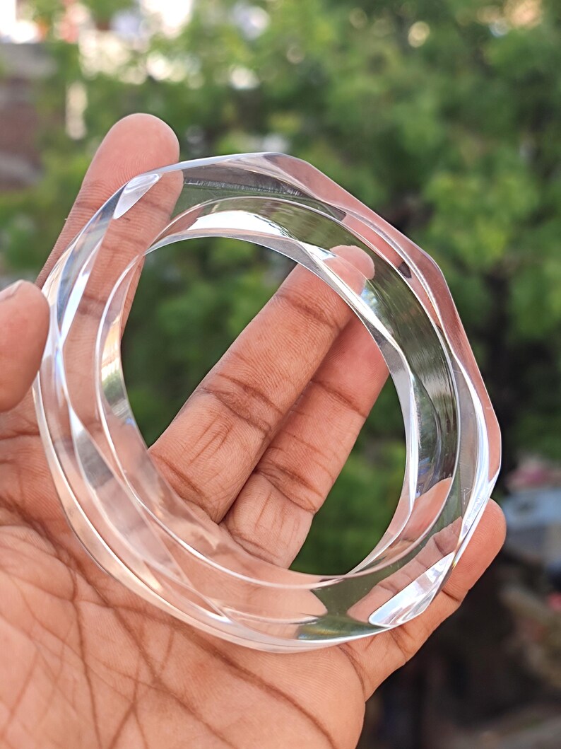 Puede incluir: Brazalete de acr&iacute;lico transparente y facetado. El brazalete transparente tiene un dise&ntilde;o geom&eacute;trico con m&uacute;ltiples lados. La luz atrapa los bordes, creando reflejos. El brazalete se sostiene en una mano sobre un fondo verde borroso.