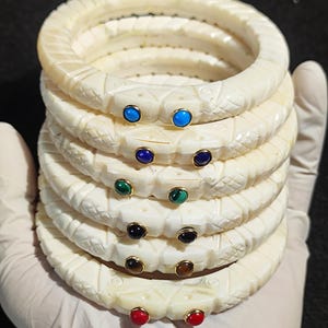 May include: A stack of six white bone bangles with intricate carvings. Each bangle has two small gemstone accents, featuring a variety of colours including turquoise, blue, green, black, brown, and red.