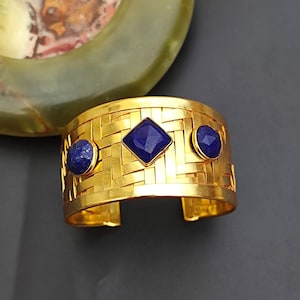 以下が含まれることがあります： 織り模様と3つの青い宝石のアクセントが付いたゴールドのカフブレスレット