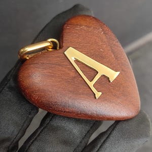 May include: A heart-shaped pendant crafted from dark brown wood, featuring a polished gold-tone metal letter A centered on the front surface. The pendant includes a gold-tone metal bail at the top for attachment to a chain or cord, showcasing a smooth finish.