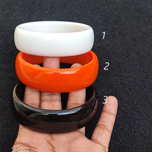 May include: Three plastic bangles stacked on top of each other. The top bangle is white, the middle bangle is orange, and the bottom bangle is black.