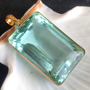 Puede incluir: Un colgante rectangular con una gran piedra preciosa azul claro engastada en un marco dorado. El colgante tiene un lazo en la parte superior para una cadena. La piedra preciosa está facetada, lo que le da un aspecto brillante. El colgante se muestra sobre una superficie blanca.