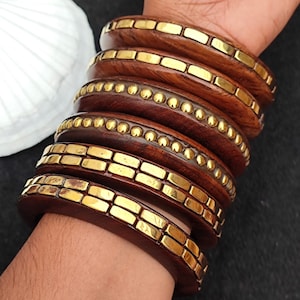 Puede incluir: Un conjunto de ocho brazaletes de madera con detalles dorados. Los brazaletes están apilados en la muñeca de una persona. Los brazaletes tienen diferentes diseños, incluyendo un patrón geométrico y un patrón de puntos.