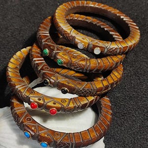 May include: Five intricately carved wooden bangles with colourful gemstone accents. The bangles are stacked on top of each other, with the top bangle featuring a white gemstone, the next bangle featuring a green gemstone, the next bangle featuring a black gemstone, the next bangle featuring a red gemstone, and the bottom bangle featuring a blue gemstone.