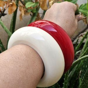 May include: Two bangle bracelets, one white and one red, worn on a person's wrist.