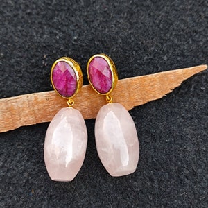 Op de afbeelding: Een paar oorhangers met goudkleurige zettingen. Elke oorbel heeft een felroze robijn bovenop en een grote, lichtroze, ovaalvormige steen eronder. De oorbellen worden gepresenteerd op een houten oppervlak tegen een donkere achtergrond.