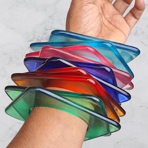 May include: A stack of colorful, translucent plastic bangles in shades of green, blue, red, pink, and purple. The bangles are stacked on a person's arm.