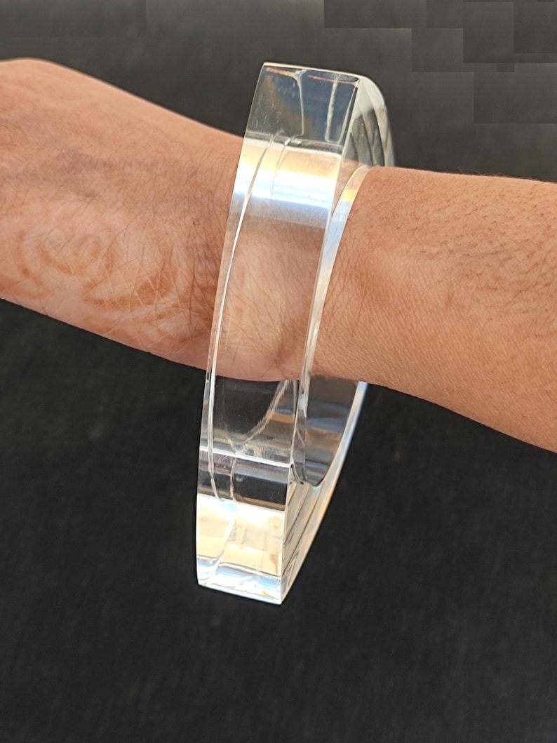 Op de afbeelding: Een heldere, rechthoekige acrylarmband. De armband is transparant en heeft een glad, gepolijst oppervlak. Het ontwerp is eenvoudig en modern, geschikt voor dagelijks gebruik. De armband is ongeveer 6,35 cm breed.